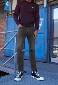 Personne portant un sweat à capuche violet foncé, un jean gris et des baskets noires montantes debout près de portes métalliques bleues numérotées 4D et 7.