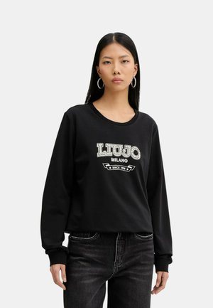 Donna con lunghi capelli neri che indossa orecchini a cerchio d'argento, felpa nera con scritta "LIU JO MILANO SINCE 1995" e jeans scuri, in piedi davanti a uno sfondo bianco.