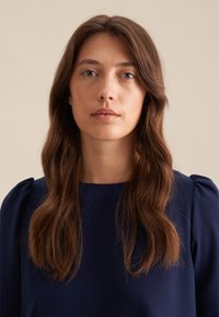 Blusa a maniche lunghe blu navy con dettagli sulle spalle a sbuffo; tessuto morbido e scollo rotondo. Modello con capelli ondulati di lunghezza media.