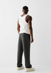 Pantalon de survêtement gris avec poches et coupe ajustée, associé à un débardeur blanc. La personne se tient sur un fond neutre.