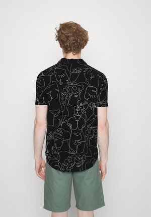 Homme aux cheveux bouclés portant un t-shirt noir à manches courtes avec des visages abstraits blancs en ligne continue et un short vert, debout face à un mur blanc.