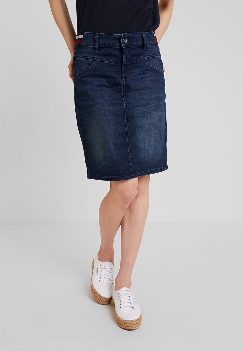 Falda lápiz de mezclilla azul oscura con diseño ajustado, bolsillos frontales y cierre de botón. Combinada con zapatillas de esparto blancas.