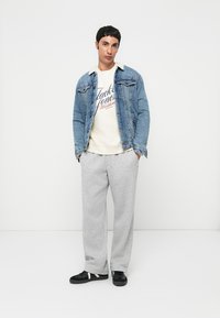 Jeansjacka över en krämfärgad sweatshirt med tryckt text, ihopparat med ljusgrå sweatpants och svarta sneakers med vita detaljer.