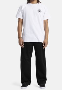 Bílé bavlněné tričko s krátkými rukávy a černým kulatým logem na hrudi, doplněné volnými černými kalhotami a černými teniskami.