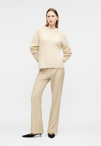 Femme portant un pull en maille beige et un pantalon large assorti avec des détails à boutons, posant les mains derrière le dos et des talons noirs à bout pointu.