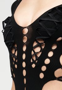 Vêtement en maille noire avec des découpes circulaires symétriques et des rubans croisés laissant apparaître la peau en dessous.