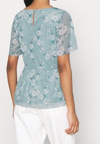Světle modrá blůza s květinovým vzorem, krátké rukávy, peplum pas a odpovídající knoflíková klíčová dírka vzadu. Průhledná textura.