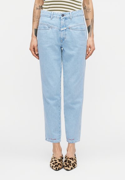 Personne portant un jean taille haute bleu clair et des chaussons à imprimé léopard, debout devant un fond blanc uni.
