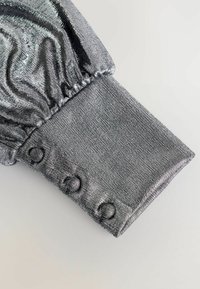 Metalická strieborná tkanina s textúrovaným povrchom, ktorá má manžetu s tlačidlami a elastickým zberom na otvorení rukáva.