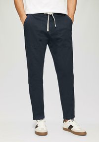 Man in marineblauwe broek met koordjes en voorzakken, met witte sneakers met zwarte strepen, staande met zijn handen in zijn zakken.