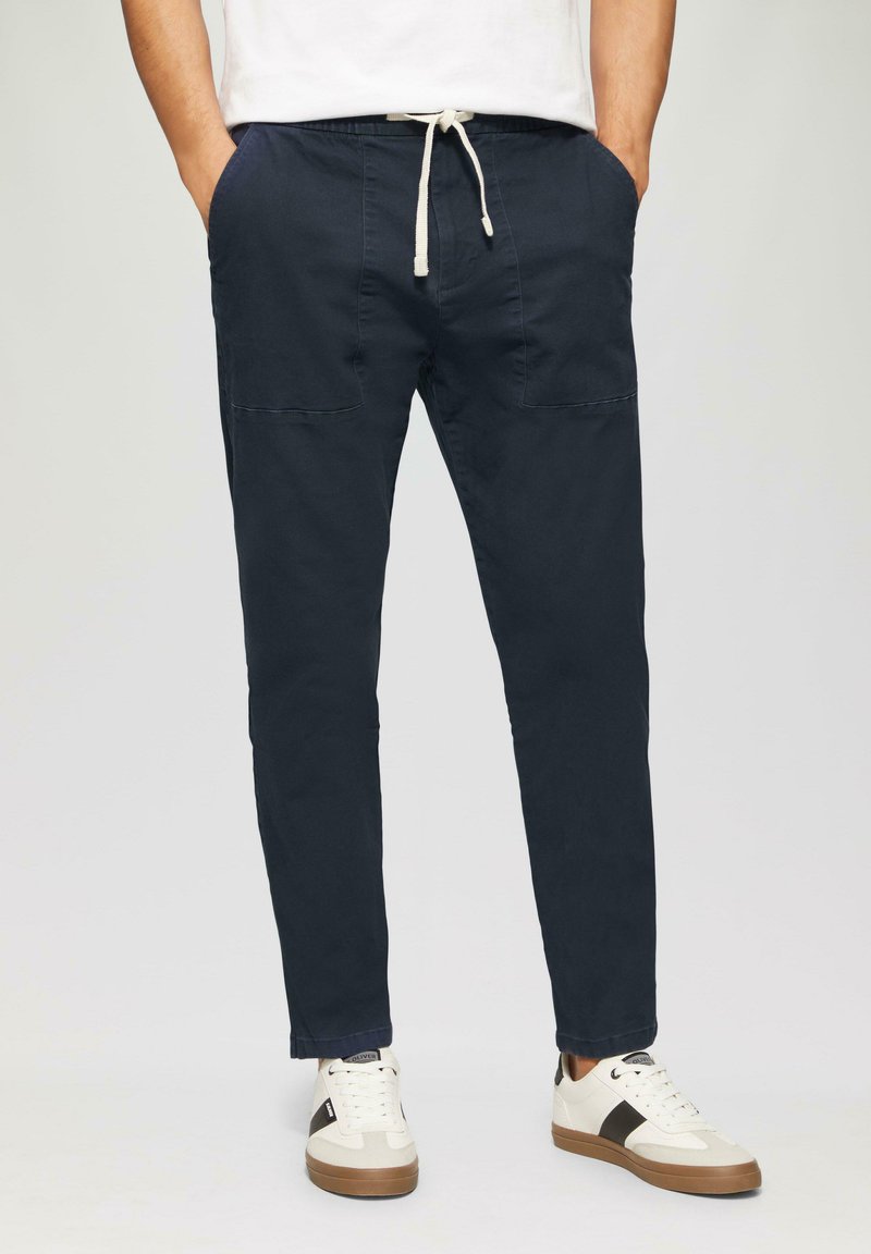 Man in marineblauwe broek met koordjes en voorzakken, met witte sneakers met zwarte strepen, staande met zijn handen in zijn zakken.