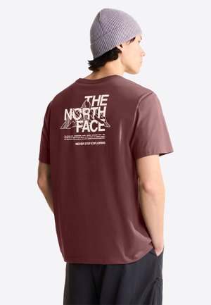 Mężczyzna w bordowej koszulce The North Face i szarej czapce, stoi z rękami w kieszeniach, bokiem pokazując logo i napis na plecach.