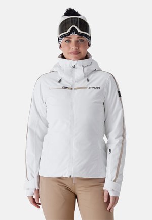 Femme portant une veste de ski blanche, un pantalon beige, un bonnet à motif noir et blanc, et un masque de ski sur le front, face à un fond neutre.