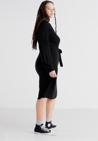 Vestido negro de punto con mangas largas y cinturón en la cintura, que presenta una silueta ajustada y un dobladillo hasta la rodilla, combinado con zapatillas altas negras.
