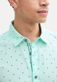 Mintgroene button-up shirt met kleine zwarte driehoekige patronen, ontworpen met een kraag en zichtbare contrasterende stiksels langs de knoopsluiting.