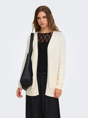 Jonge vrouw met golvend haar, een crème cardigan over een zwarte kanten top en zwarte broek dragend, met een grote zwarte schoudertas.