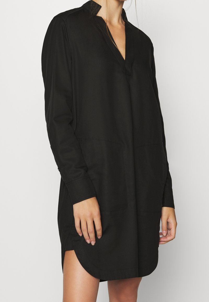 Femme portant une robe chemise noire ample à manches longues avec un col en V, debout devant un fond clair uni.