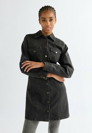 Young woman with braided hair wears a black long-sleeve buttoned shirt dress with a subtle reptile pattern, posing with arms crossed.