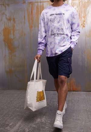 Person i lilla tie-dye langærmet trøje og marineblå cargoshorts går, mens de bærer en beige tote-taske med gul blokdesign.