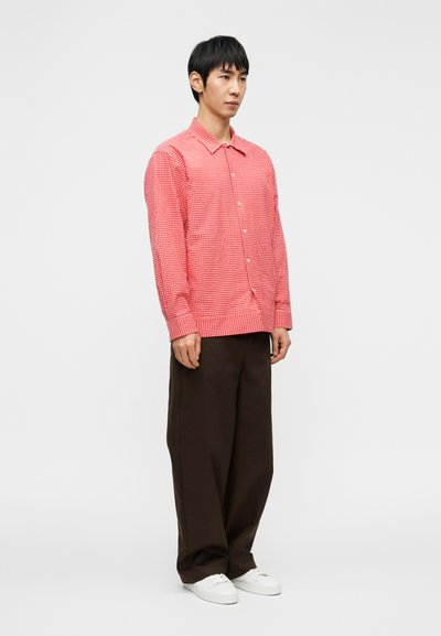 Κόκκινο και άσπρο πουκάμισο με πουά σχέδιο και κουμπιά, μακριά μανίκια, φορεμένο με άνετο καφέ παντελόνι και λευκά αθλητικά παπούτσια. Απλός γιακάς, ορατά κουμπιά στο μπροστινό μέρος.