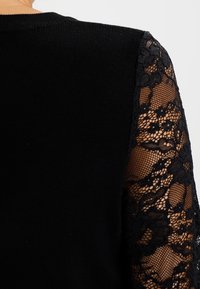 Haut noir en tissu côtelé solide sur le corps et en dentelle noire transparente sur les manches, avec des motifs floraux et des designs texturés.