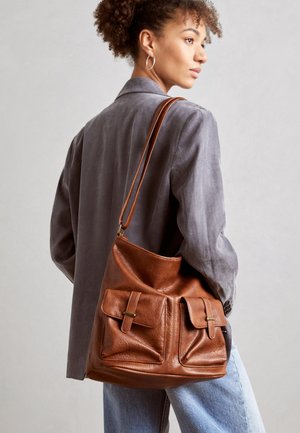 Woman wearing gray blazer and light blue jeans carries large brown leather shoulder bag with front pockets.