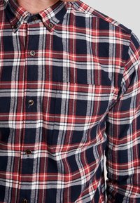 Camicia a quadri rossa, blu navy e bianca con bottoni. Presenta un colletto, una tasca sul petto e accenti neri in contrasto sui bottoni frontali. Materiale in cotone.