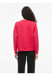 Rød genser i mykt materiale med avslappet passform, senkede skuldre og ribbestrikkede mansjetter. Kombinert med svarte bukser, sett bakfra.