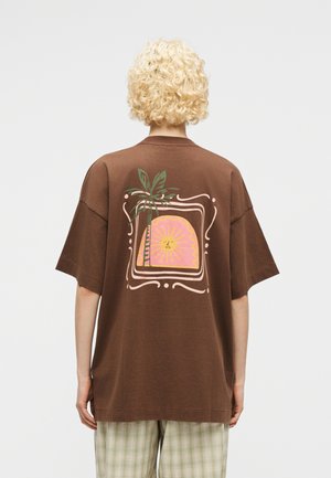 Personne aux cheveux blonds bouclés portant un T-shirt marron oversized avec un graphisme de soleil et de palmier dans le dos et un pantalon à carreaux beige.