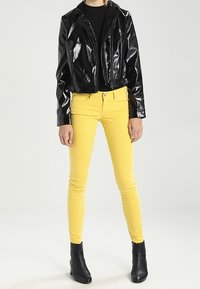 Chaqueta de motorista negra y brillante, corta, sobre un top negro, combinada con jeans ajustados amarillos brillantes y botines negros. Texturas suaves en toda la prenda.