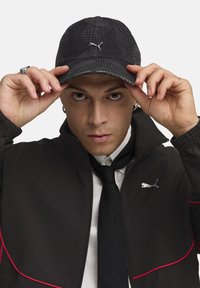Sort cap med en struktureret overflade og sølv Puma-logo, båret af en person i en sort jakke med røde accenter og et sort slips.