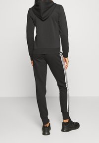 Sweat à capuche noir, associé à un jogging noir avec des rayures blanches sur les côtés. Le matériau semble doux ; design ajusté avec des poignets en côtes.
