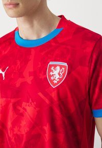 Rød fodboldtrøje med blå krave, der har et struktureret mønster og et hvidt løvemblem på et skjold. Lavet af åndbart stof.