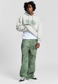 Felpa grigia con un design grafico verde, indossata sopra una maglietta bianca; abbinata a pantaloni larghi verdi a trama e sneakers nere.