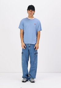 Camisa de manga corta azul claro con un cuello en contraste rojo y blanco, combinada con pantalones cargo azules de corte holgado y zapatillas grises. Gorra negra.
