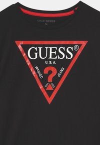 Svart bomullströja med en stor röd triangel-logga som har texten "GUESS" och "WASHED JEANS", vita accenter och en särskild utformning av ett frågetecken.