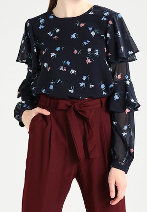 Kobieta ubrana w granatową kwiecistą bluzkę z warstwowymi, falbanianymi rękawami oraz bordowe spodnie z wysokim stanem i przewiązanym materiałowym paskiem.
