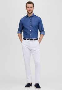 Camicia in denim a maniche lunghe di un blu scuro, pantaloni bianchi, cintura nera e scarpe slip-on di color navy. Il modello posa con le mani in tasca su uno sfondo neutro.