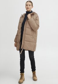 Beige padded coat with a hood, front zipper, and side pockets. Worn over a dark sweater with black jeans and tan lace-up boots.