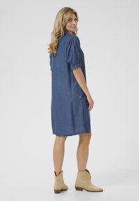 Rochie din denim albastru, cu un croi lejer, mâneci scurte și bufante, și buzunare laterale. Prezintă un tiv care ajunge deasupra genunchiului, purtată cu cizme bej.