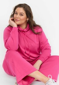 Felpa fucsia con cappuccio e coulisse, abbinata a pantaloni a gamba larga coordinati. Tessuto morbido e vestibilità rilassata, indossata con sneakers bianche.