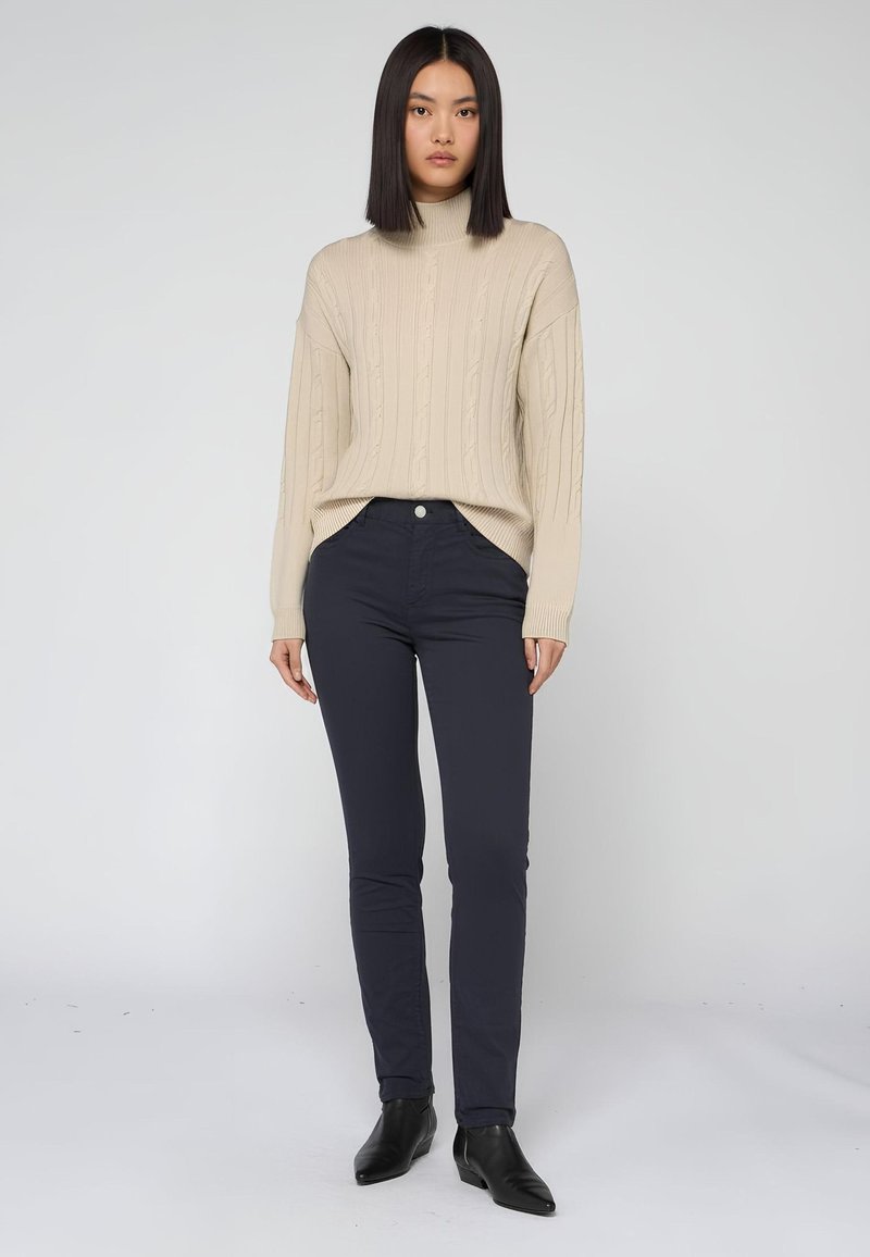 Beige kabelgebreide trui met hoge nek, gecombineerd met donkerblauwe nauwsluitende pants en zwarte enkellaarzen. Soepele texturen en eenvoudig ontwerp.