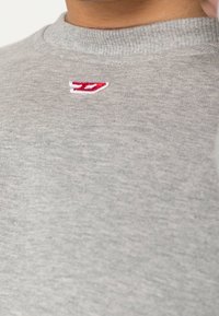 Šedá mikina s kulatým výstřihem a měkkou strukturou, s malým červeným a bílým vyšitým logem na horní levé části hrudi.