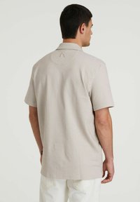 Camisa de manga corta con cuello en tono beige claro y sutiles rayas verticales. Presenta un dobladillo redondeado y un diseño circular en el cuello de la parte posterior.