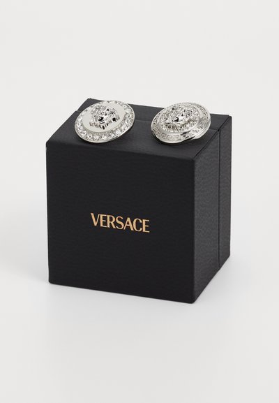 Stříbrné kulaté náušnice s detailem lva, obklopené čirými krystaly, ležící na černé krabičce Versace se zlatým logem.