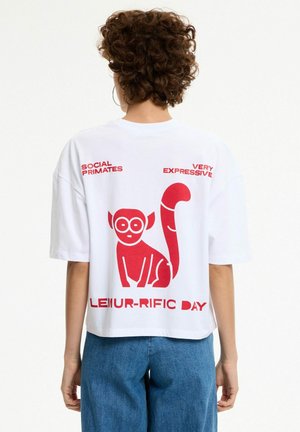 Persona con cabello rizado lleva puesta una camiseta blanca con un lémur de dibujos animados rojo y el texto "Social Primates," "Very Expressive," y "Lemur-rific Day" en la espalda.