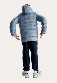 Person som bär en ljusblå puffjacka med huva, mörka byxor, vita strumpor och grå sneakers, stående med armarna böjda bakåt.