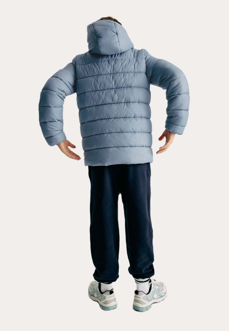 Person som bär en ljusblå puffjacka med huva, mörka byxor, vita strumpor och grå sneakers, stående med armarna böjda bakåt.