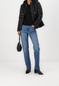 Zwarte gewatteerde jas met een capuchon, zwarte top, blauwe rechte spijkerbroek, en puntige zwarte hoge hakken. Houdt een kleine zwarte tas vast.