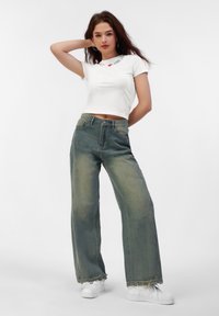 Hvid cropped t-shirt med teksten "I ❤️ Liebe". Løs-fittede, falmede grønne jeans med frayed kanter. Model iført hvide sneakers.